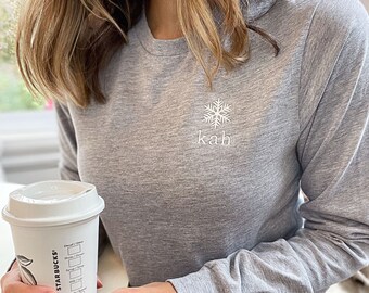 May include: A gray long-sleeved shirt with a white snowflake and the word "kah" embroidered on the front. The shirt is being worn by a person holding a cup of coffee.
