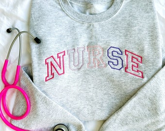 May include: Gray sweatshirt with the word "NURSE" embroidered in rainbow colors. A pink stethoscope is lying on top of the sweatshirt.