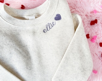 May include: A cream-coloured sweatshirt with a purple heart and the name "Ellie" embroidered on the front.