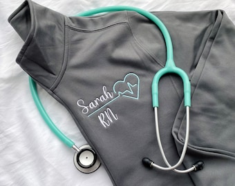 Puede incluir: Sudadera gris con un corazón bordado en blanco y el texto "Sarah RN". Un estetoscopio turquesa está encima de la sudadera.