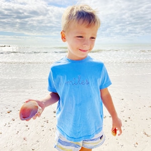 May include: A young boy wearing a light blue t-shirt with the word "miles" embroidered on the front. He is standing on a sandy beach and holding a seashell.