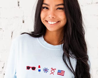 May include: Light blue t-shirt with embroidered patriotic icons: sunglasses, lipstick, a firework, a bow, and an American flag. The shirt is made of a soft material and has a crew neck.