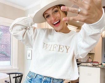 Op de afbeelding: Een vrouw draagt een wit sweatshirt met het woord "WIFEY" geborduurd op de voorkant, een blauwe denim short en een witte fedora. Ze glimlacht en laat een diamanten verlovingsring zien aan haar linkerhand.