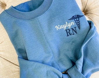 Puede incluir: Una sudadera azul con el texto "Kaylynn RN" y un símbolo médico bordado en la parte delantera.