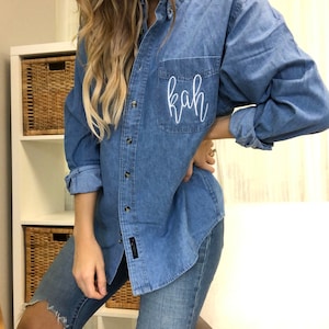 May include: A light blue denim shirt with the initials "kah" embroidered on the chest pocket. The long-sleeved shirt has button closures and a classic collar. The sleeves are rolled up. The shirt is paired with blue jeans.