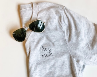 May include: Gray t-shirt with the text "dog mom" embroidered on the front. Sunglasses are lying on top of the shirt.