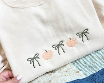 Puede incluir: Sudadera de color crema con bordados de calabazas y lazos. El diseño presenta tres calabazas color melocotón y cuatro lazos verdes. La sudadera está doblada sobre una tela estampada azul y blanca.