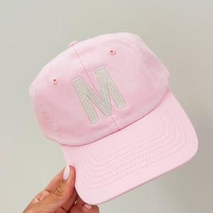 May include: A light pink baseball cap with a curved brim. The cap features a large, embroidered letter "M" in a lighter shade of pink on the front. The cap is made of a soft fabric.