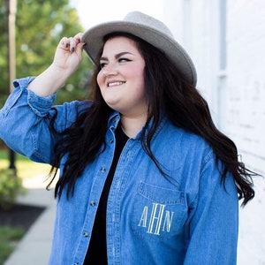 May include: A denim shirt with a pocket featuring the embroidered initials "AHN" in white. The shirt is buttoned and worn with a gray hat. The person is smiling and looking to the side.