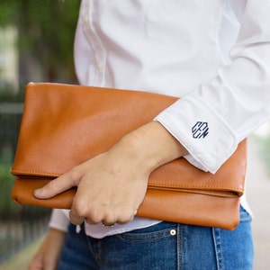 May include: A white button-down shirt with long sleeves and a monogram on the cuff. The shirt is tucked into blue jeans and the person is holding a brown leather clutch.