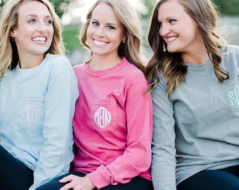 May include: Three women wearing long-sleeved shirts with embroidered monograms on the pocket. The shirts are light blue, pink, and gray. The women are sitting on a stone wall.