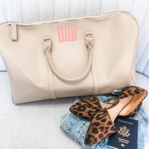 May include: A beige travel bag with a pink monogram, leopard print flats, a denim jacket, and a United States passport. The bag has a zip closure and two handles.
