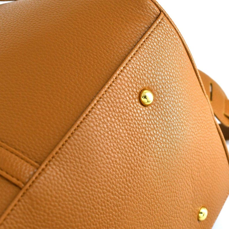 May include: Close-up of a tan leather handbag. The bag features a textured surface, visible stitching, and gold-coloured metal studs. The bag's design includes a structured shape and a strap.
