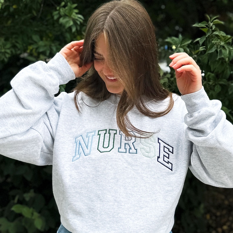 young woman wearing an ash crewneck sweatshirt with custom NURSE embroidered design in shades of blue and green