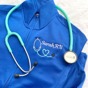 May include: Blue fleece jacket with a stethoscope embroidered on the front. The text "Sarah, RN" is embroidered below the stethoscope.