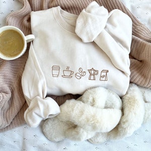 Op de afbeelding: Een wit sweatshirt met een bruine afbeelding van koffiekopjes, koffiebonen en een koffiekan. Het sweatshirt ligt op een pluche witte deken met een paar witte pluche pantoffels.