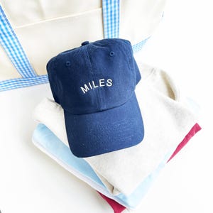 May include: A navy blue baseball cap with the name "MILES" embroidered in white. The cap sits atop a stack of folded shirts in light blue, beige, and burgundy. A white tote bag with a blue gingham trim is in the background.