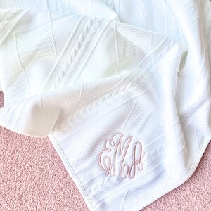 May include: White cable knit baby blanket with a pink embroidered monogram "EM"