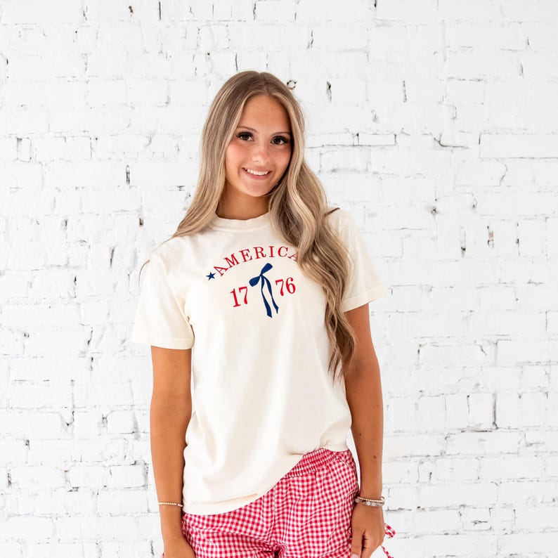 Girl standing in front of a white brick wall wearing an ivory comfort colors t-shirt and red and white gingham shorts. Tank top features a custom embroidered design of America arched over a long tail ribbon bow framed by 1776. Embroidery is done in red and navy thread.