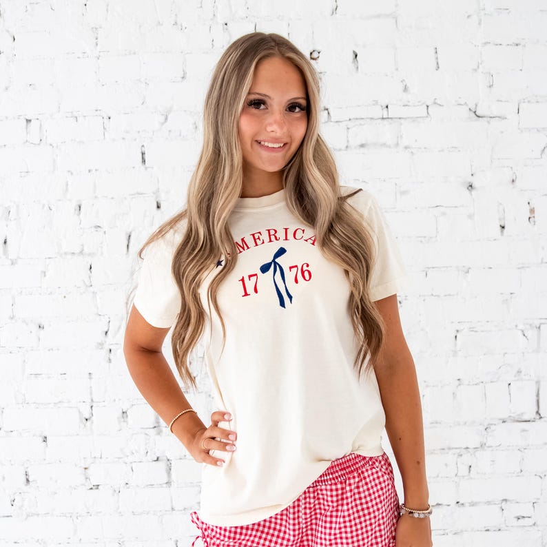 Girl standing in front of a white brick wall wearing an ivory comfort colors t-shirt and red and white gingham shorts. Tank top features a custom embroidered design of America arched over a long tail ribbon bow framed by 1776. Embroidery is done in red and navy thread.