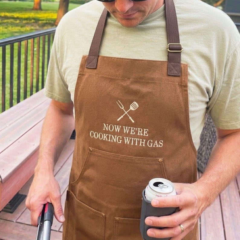 man outside grilling wearing a duck brown apron with custom embroidered text reading: "now we're cooking with gas"