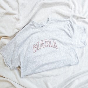 K&ouml;nnte beinhalten: Ein graues T-Shirt mit dem Wort "MAMA" in rosa auf der Vorderseite gestickt.