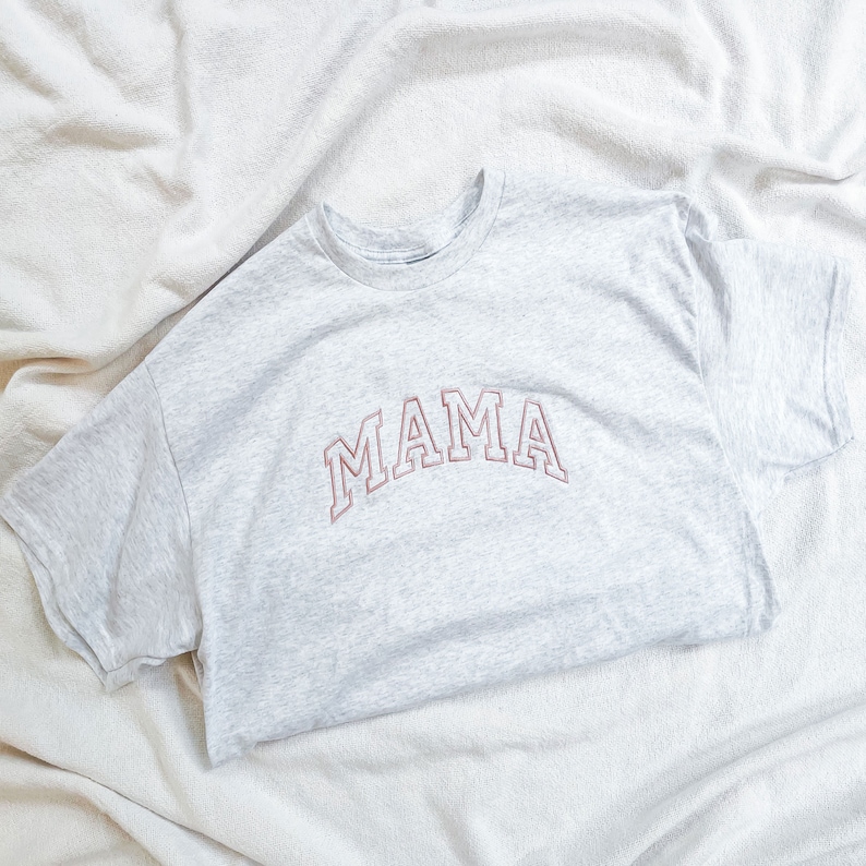 K&ouml;nnte beinhalten: Ein graues T-Shirt mit dem Wort "MAMA" in rosa auf der Vorderseite gestickt.