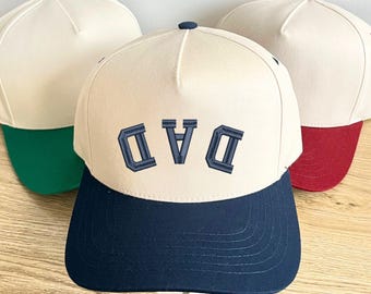 May include: Three baseball caps with a cream-colored crown and a navy blue brim. The center cap has the word "DAD" embroidered in navy blue. One cap has a green brim, and another has a red brim.