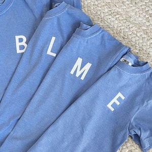May include: Four blue t-shirts stacked on top of each other. Each shirt has a white letter on the front, spelling out the word "BLME".