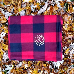 May include: A red and black buffalo plaid fleece throw blanket with a white monogram in the center.