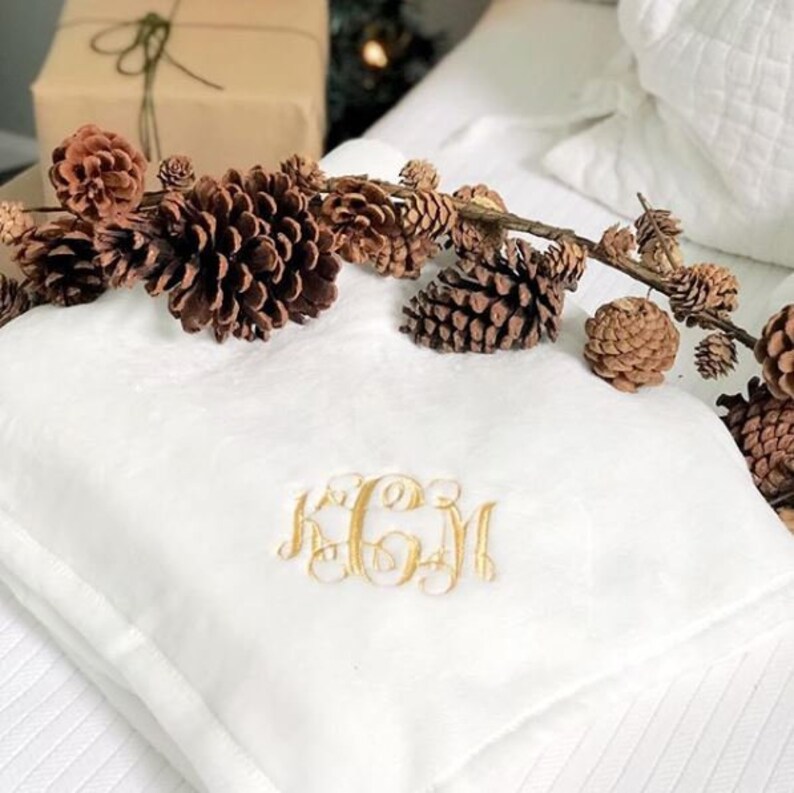 May include: White plush throw blanket with a gold embroidered monogram "KCM". The blanket is decorated with a garland of pine cones.