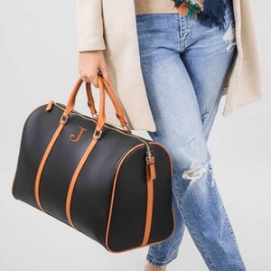 May include: Black duffel bag with tan detailing, handles, and trim. The bag has a gold zip and a gold letter "J". The bag is held by a person wearing blue jeans and a beige coat.