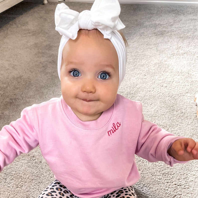 toddler girl wearing a pink crewneck sweatshirt with embroidered name along the collar