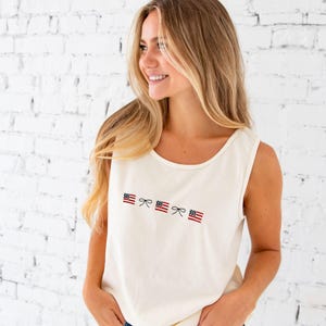 close up of a blonde woman standing in front of a white brick wall wearing denim shorts and an ivory comfort colors tank top with embroidered mini usa flags and dainty bow design across the chest in an alternating pattern