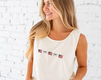 May include: Cream-colored tank top with three embroidered American flags and bows. The sleeveless top has a scoop neckline and is made of a soft, lightweight fabric. The patriotic design is perfect for celebrating Independence Day or showing national pride.