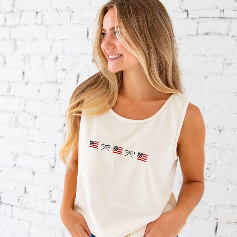 close up of a blonde woman standing in front of a white brick wall wearing denim shorts and an ivory comfort colors tank top with embroidered mini usa flags and dainty bow design across the chest in an alternating pattern