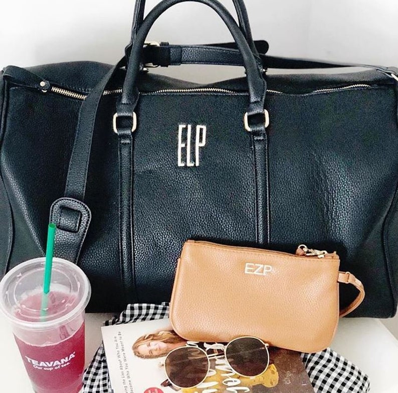 May include: Black leather holdall with gold detailing and the initials "ELP". A smaller tan clutch bag with "EZP" is next to the holdall. Sunglasses, a drink, and a book are also visible.