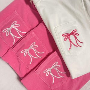 May include: Four pink t-shirts with white embroidered bows on the front pockets. The shirts are stacked on top of each other.