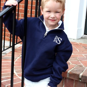May include: A young boy wearing a navy blue quarter-zip sweatshirt with white embroidered initials "B.R.S." on the chest. He is standing on a brick staircase and holding onto a black railing.