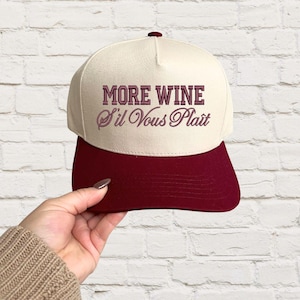 May include: A beige and burgundy baseball cap with the text "MORE WINE S'il Vous Plait" embroidered in pink.