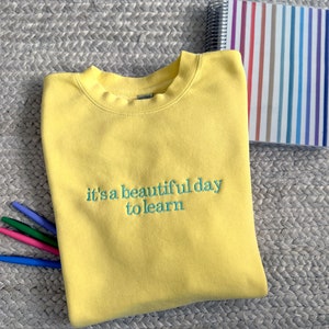 Puede incluir: Sudadera amarilla con el texto "it's a beautiful day to learn" bordado en verde.