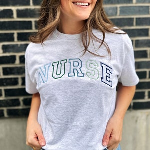 young woman wearing an ash tee with custom NURSE embroidered design in shades of blue and green