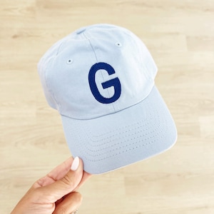 May include: A light blue baseball cap with a curved brim. The cap features a navy blue embroidered letter "G" on the front. The hat is made of a soft fabric and has a classic, casual style.
