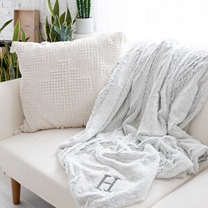 Pu&ograve; includere: Un divano bianco con un cuscino strutturato color crema e una coperta in finta pelliccia grigio chiaro. La coperta &egrave; drappeggiata sul divano e presenta una grande lettera "H" ricamata in grigio. Il cuscino ha un motivo quadrato e nappe.