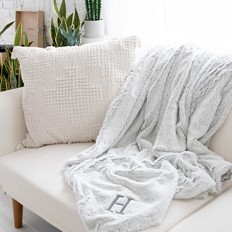 Pu&ograve; includere: Un divano bianco con un cuscino strutturato color crema e una coperta in finta pelliccia grigio chiaro. La coperta &egrave; drappeggiata sul divano e presenta una grande lettera "H" ricamata in grigio. Il cuscino ha un motivo quadrato e nappe.