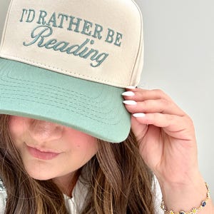 May include: A beige and mint green baseball cap with the embroidered text "I'd Rather Be Reading". The cap has a curved brim and a structured crown. The hat is a casual accessory.