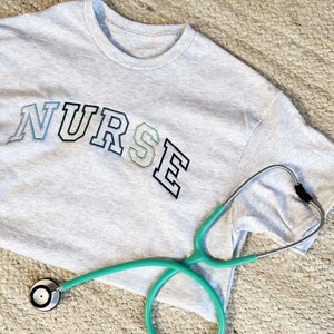 an ash crewneck tee with custom NURSE embroidered design in shades of blue and green
