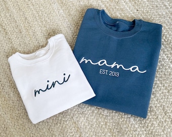 Könnte beinhalten: Zwei Sweatshirts, eines weiß und eines marineblau, werden auf einer gewebten Oberfläche gezeigt. Das weiße Sweatshirt hat das Wort "mini" in marineblauer Schrift. Das marineblaue Sweatshirt hat das Wort "mama" in weißer Schrift mit "EST. 2013" darunter.