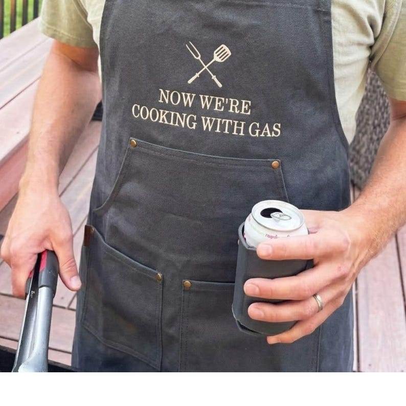 man outside grilling wearing a charcoal apron with custom embroidered text reading: "now we're cooking with gas"
