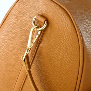 May include: Close-up of a tan leather bag with a gold-coloured metal clasp and strap attachment. The bag has a textured surface and a rounded shape, with visible stitching details.